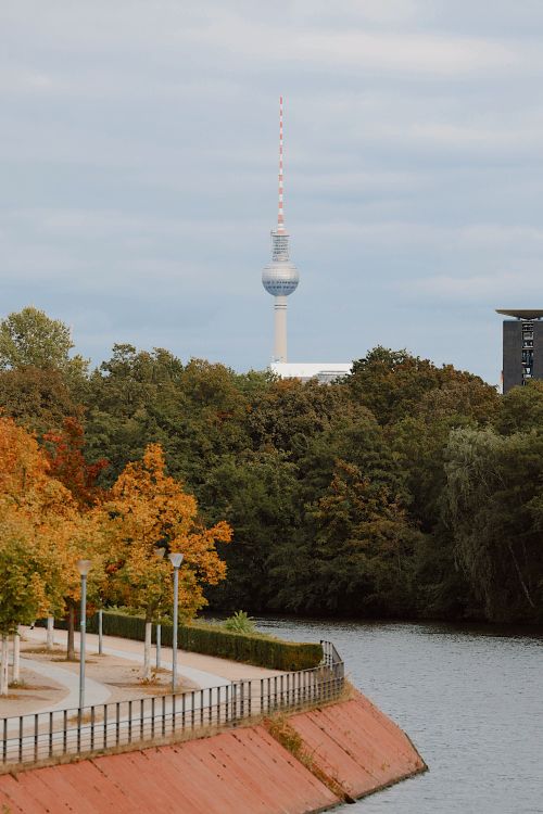Free Scenic view of Berlin TV Tower above autumn-colored trees near a riverside pathway. Stock Photo