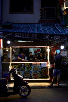 A bustling night street food stall selling sushi with neon lights and customers.