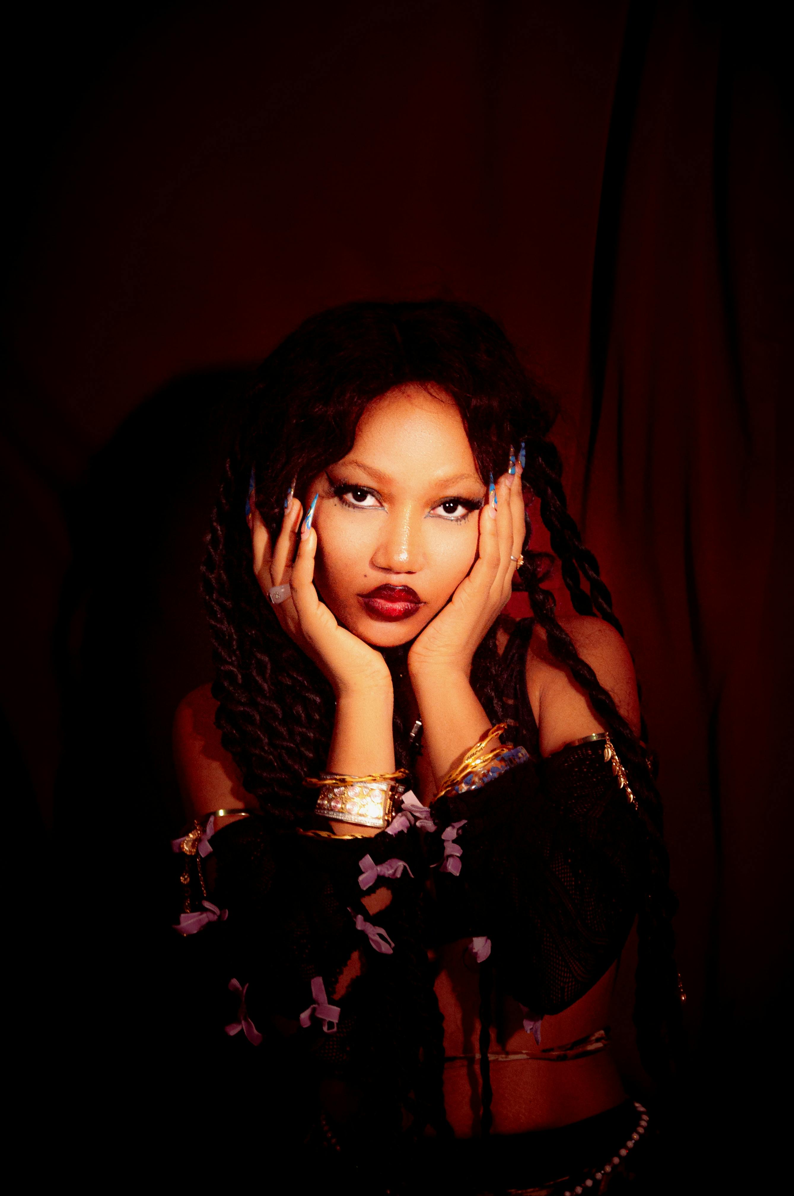 Artistic portrait of a woman with bold makeup, jewelry, and braided hair against a dark background.