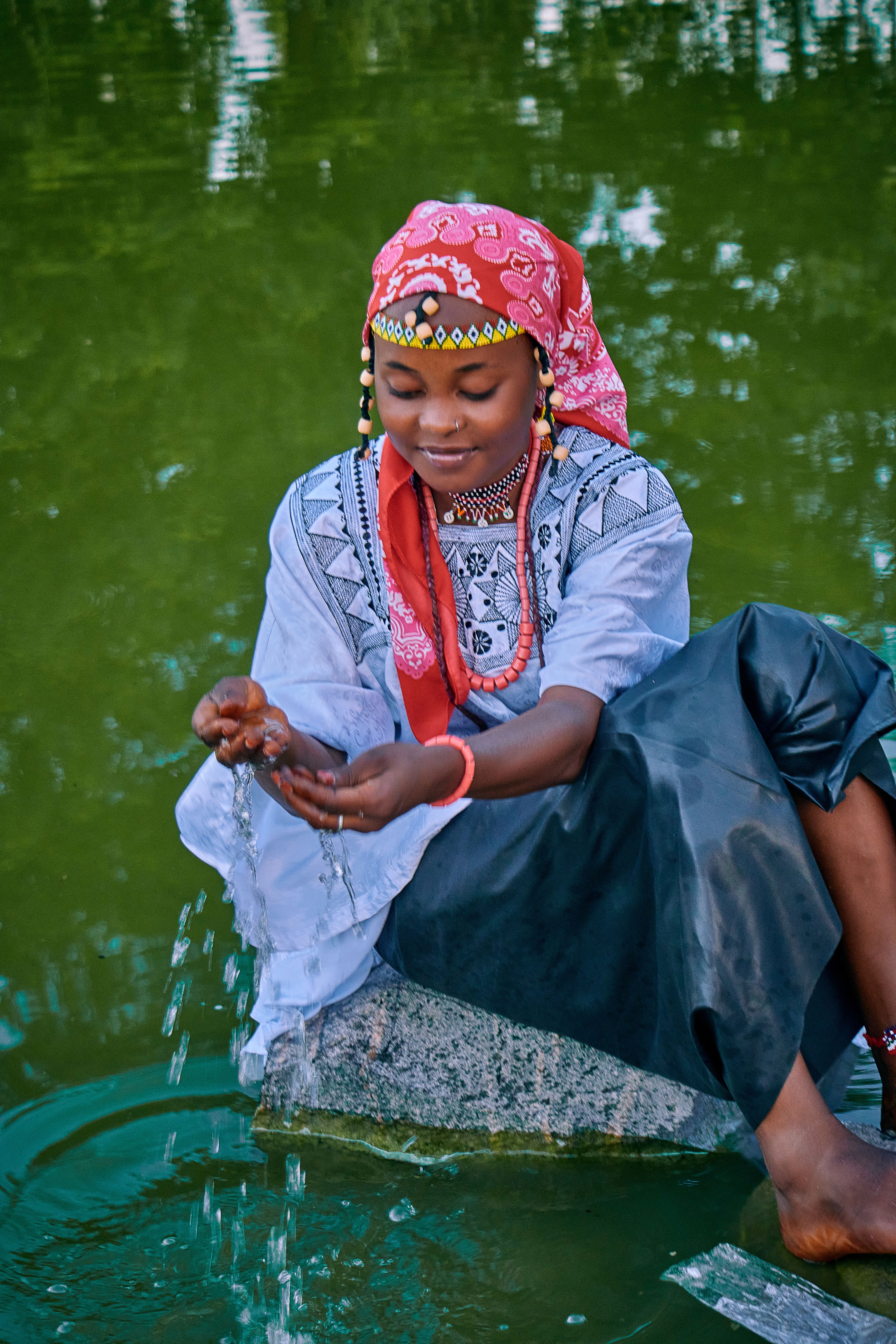 A young woman in vibrant traditional clothing sitting by a pond, splashing water.