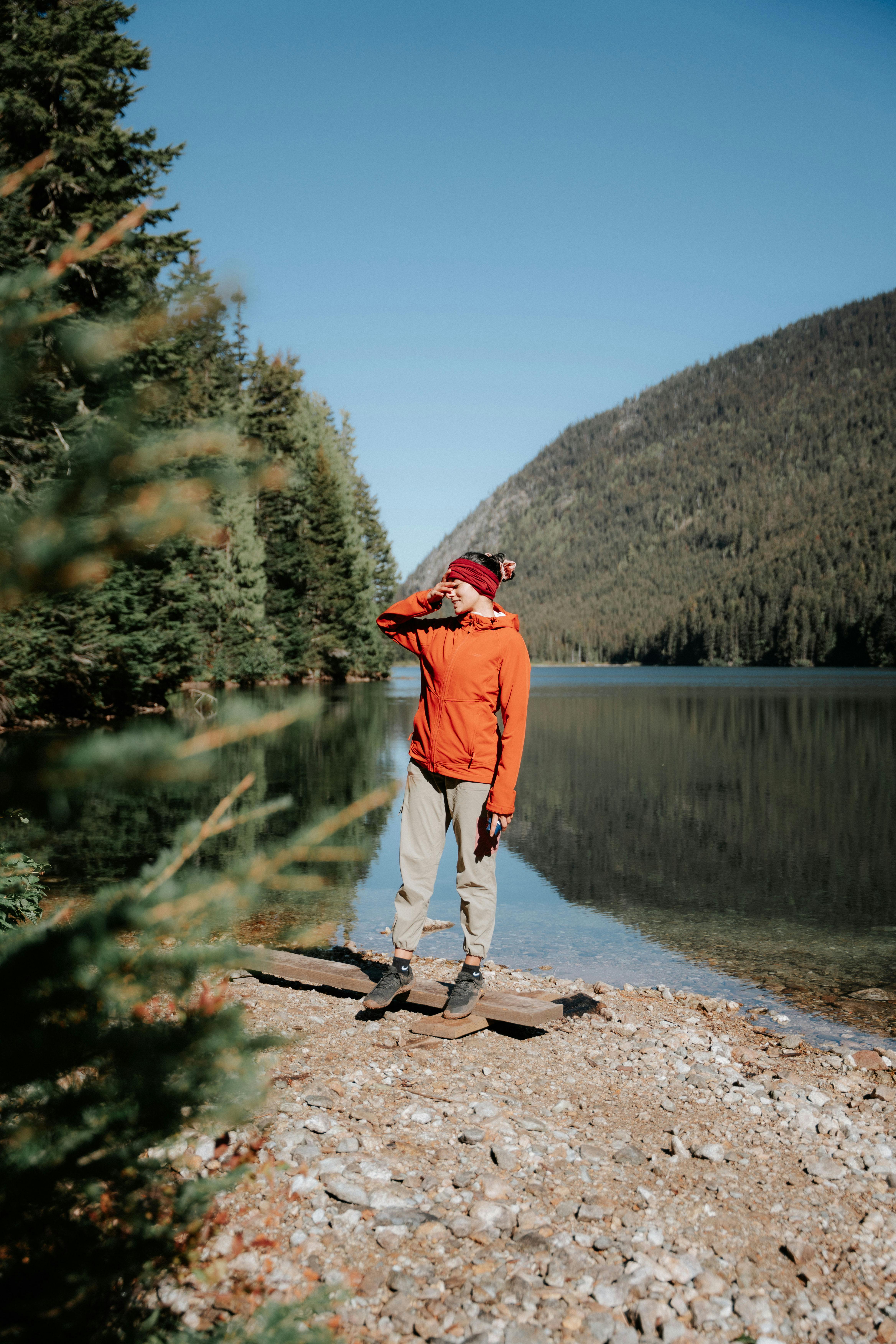 Person in orange jacket enjoys scenic lakeside view amid mountains and forest.