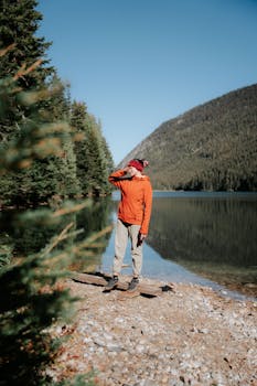 Person in orange jacket enjoys scenic lakeside view amid mountains and forest.
