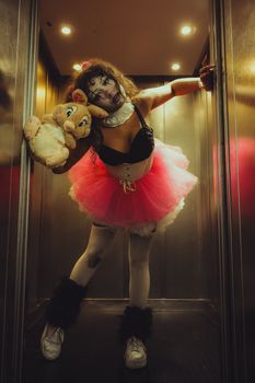 Creepy clown in a tutu with plush toy in a dimly lit elevator setting.