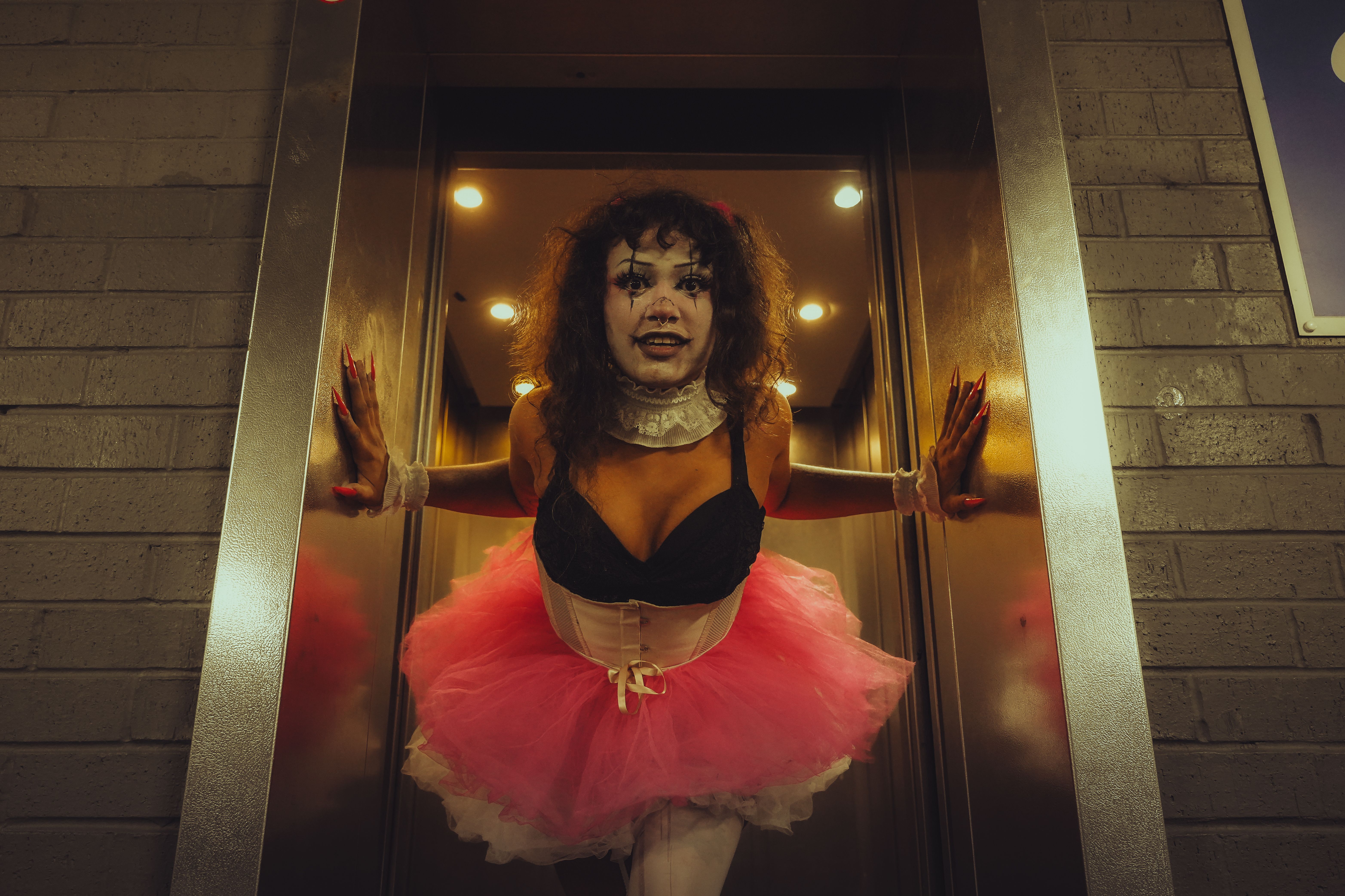 A person in clown makeup and tutu costume emerging from an elevator, showcasing gothic and horror aesthetics.