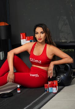 A woman in a red fitness outfit sits with nutrition supplements and a kettlebell indoors.