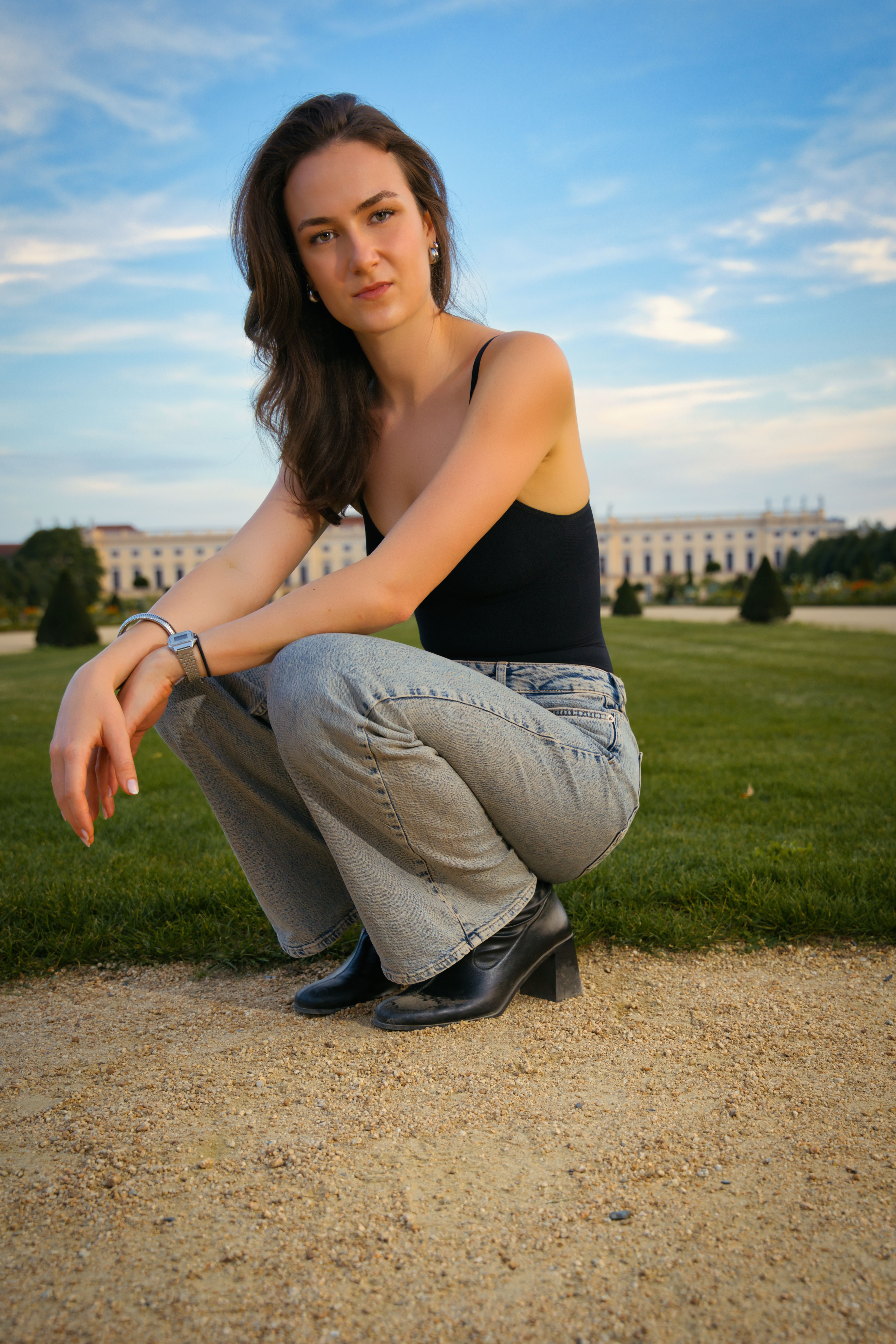 A fashionable young woman crouches on a path in a Berlin park, with a historic building in the background.