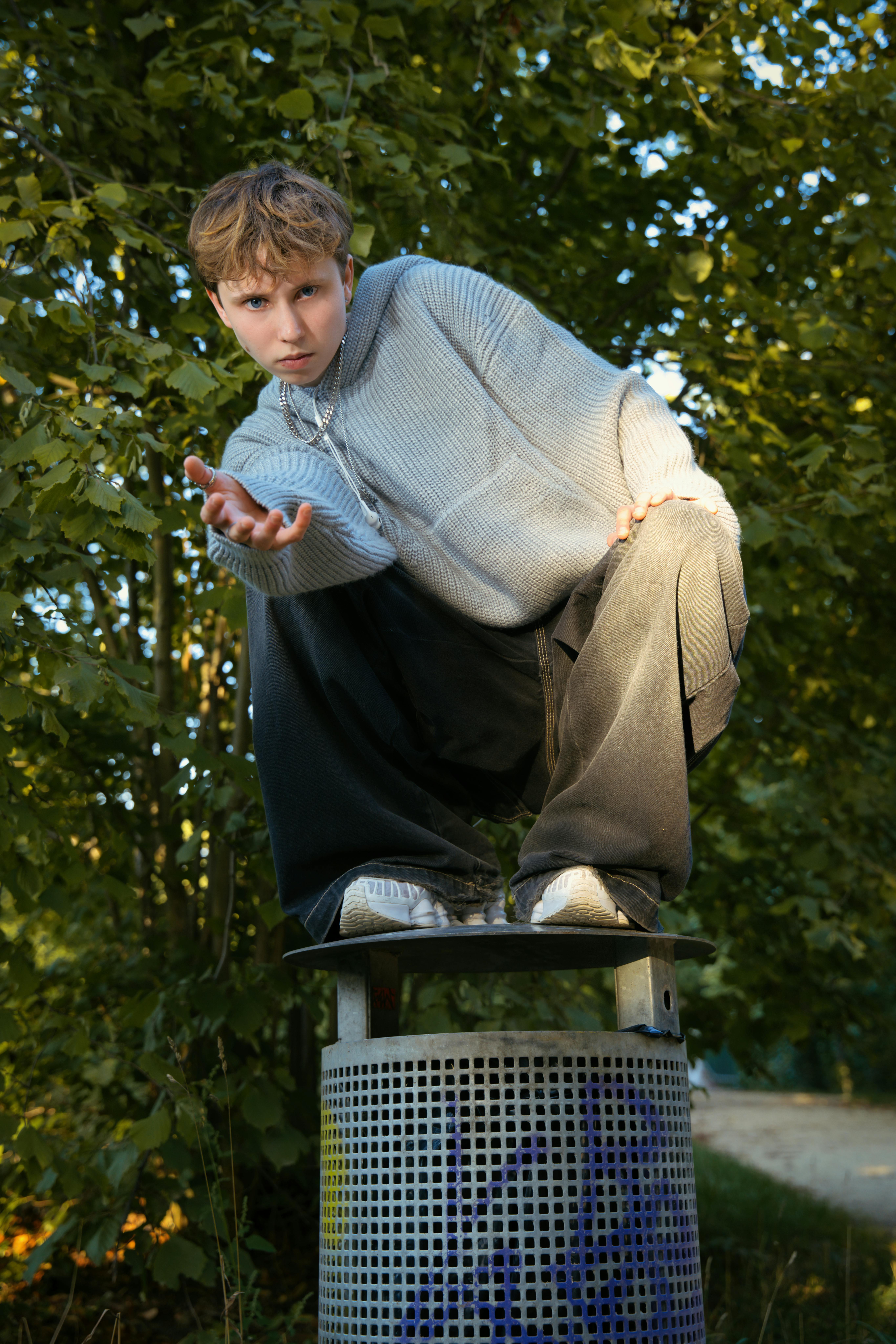 Teen crouched on a bin in a park in Berlin, surrounded by greenery.