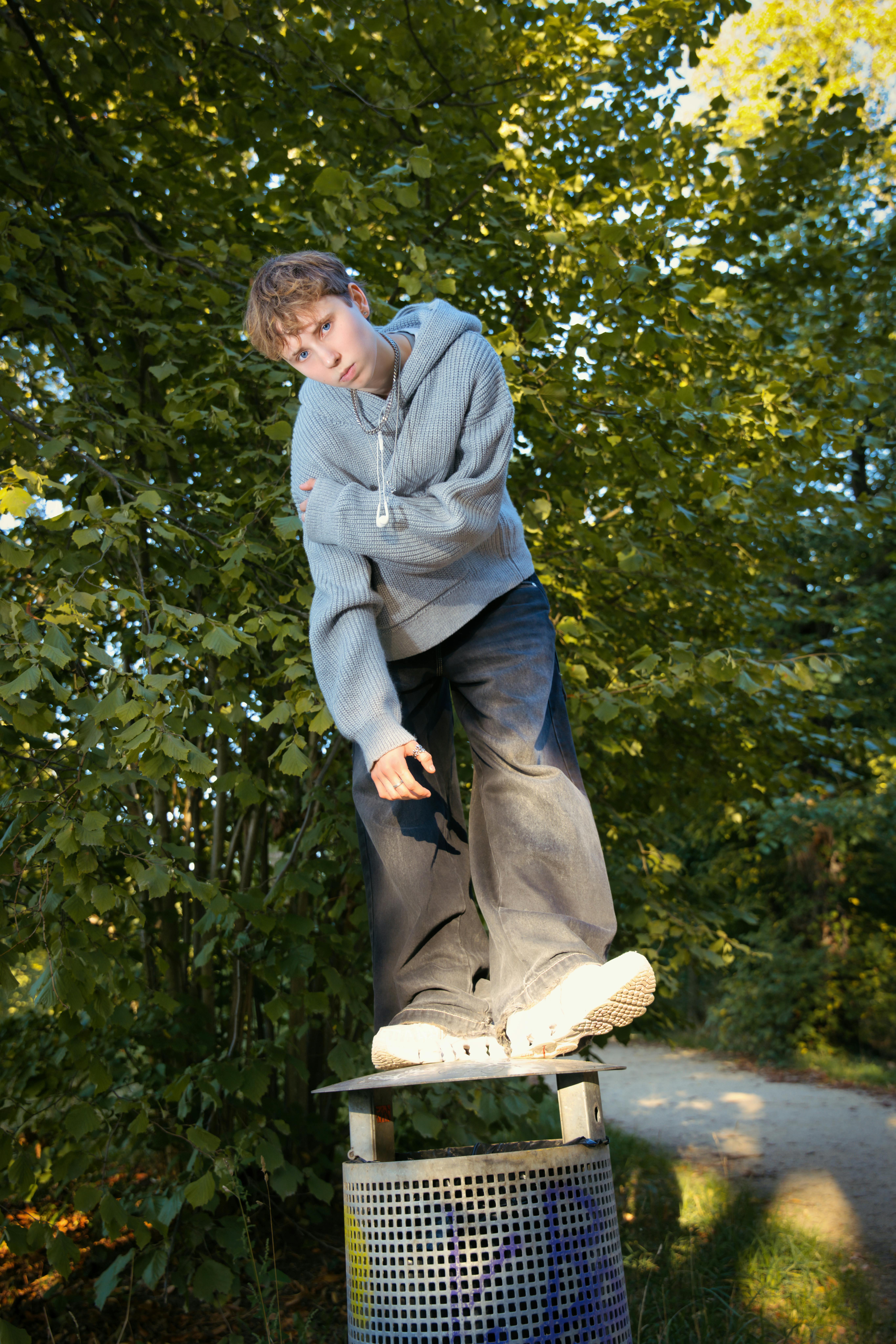 An adolescent boy skillfully balancing on a trash bin, surrounded by lush greenery in a Berlin park.