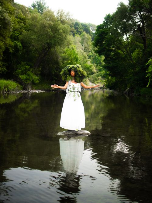 Free A woman stands on a rock in a river, surrounded by lush greenery and wearing a leafy headpiece. Stock Photo