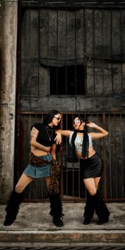 Two stylish women posing in trendy outfits against a rugged urban backdrop, exuding confidence.