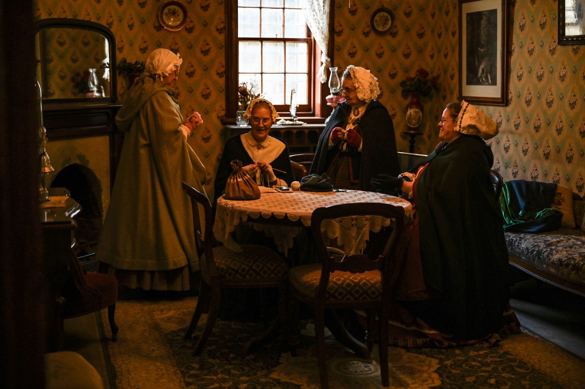 Victorian women in a cozy parlor engaged in conversation, dressed in period attire.