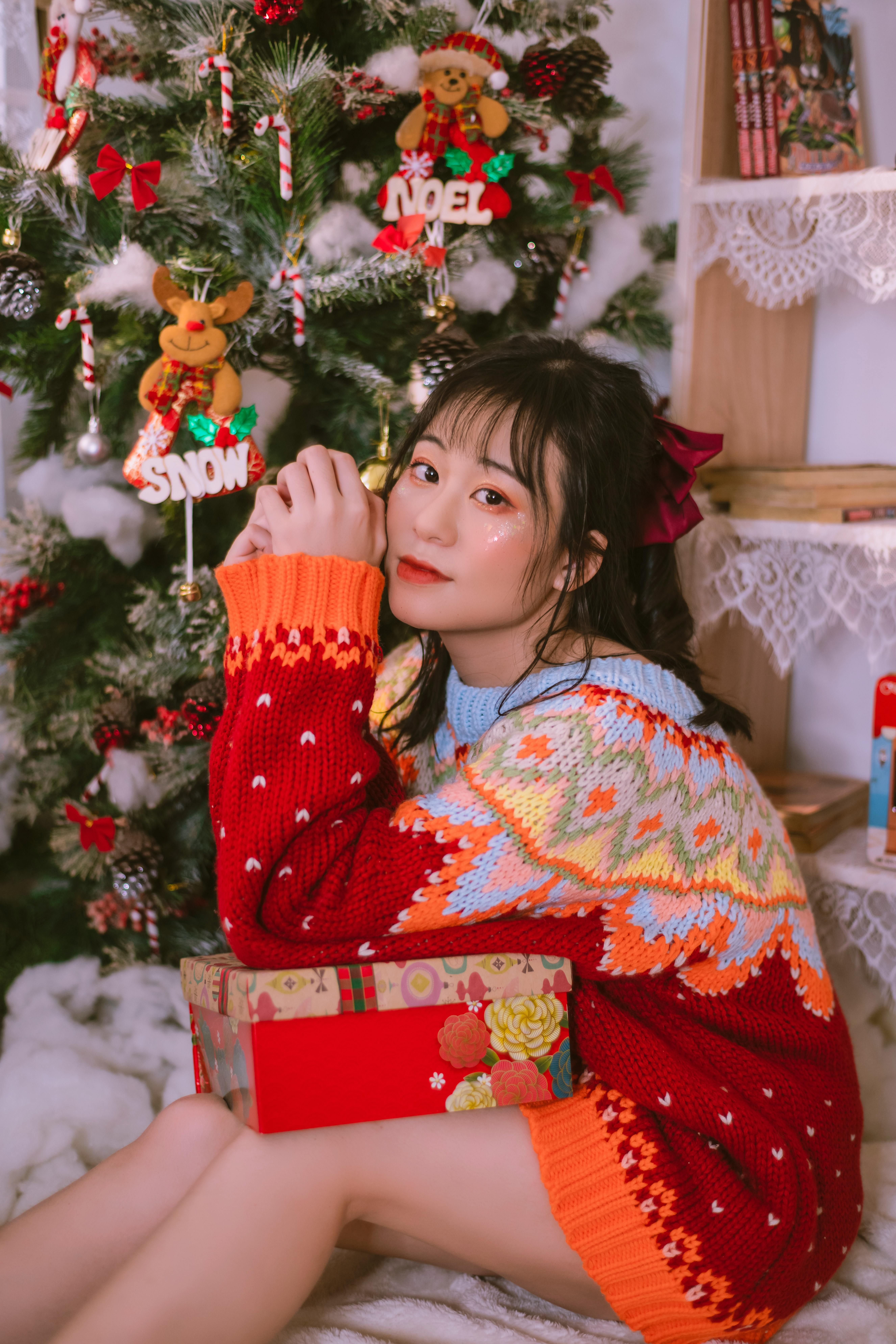 Young woman in colorful sweater sitting by a Christmas tree holding a gift.