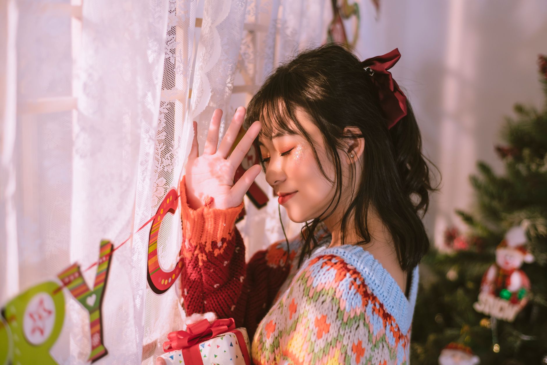 Young woman smiling by a festive window with holiday decor and gift in hand.
