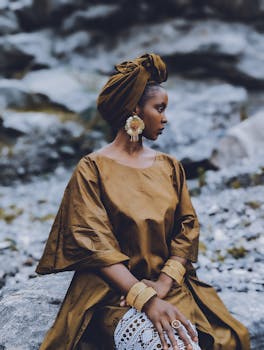Stylish woman in rich brown traditional Nigerian dress sitting outdoors on rocky terrain.