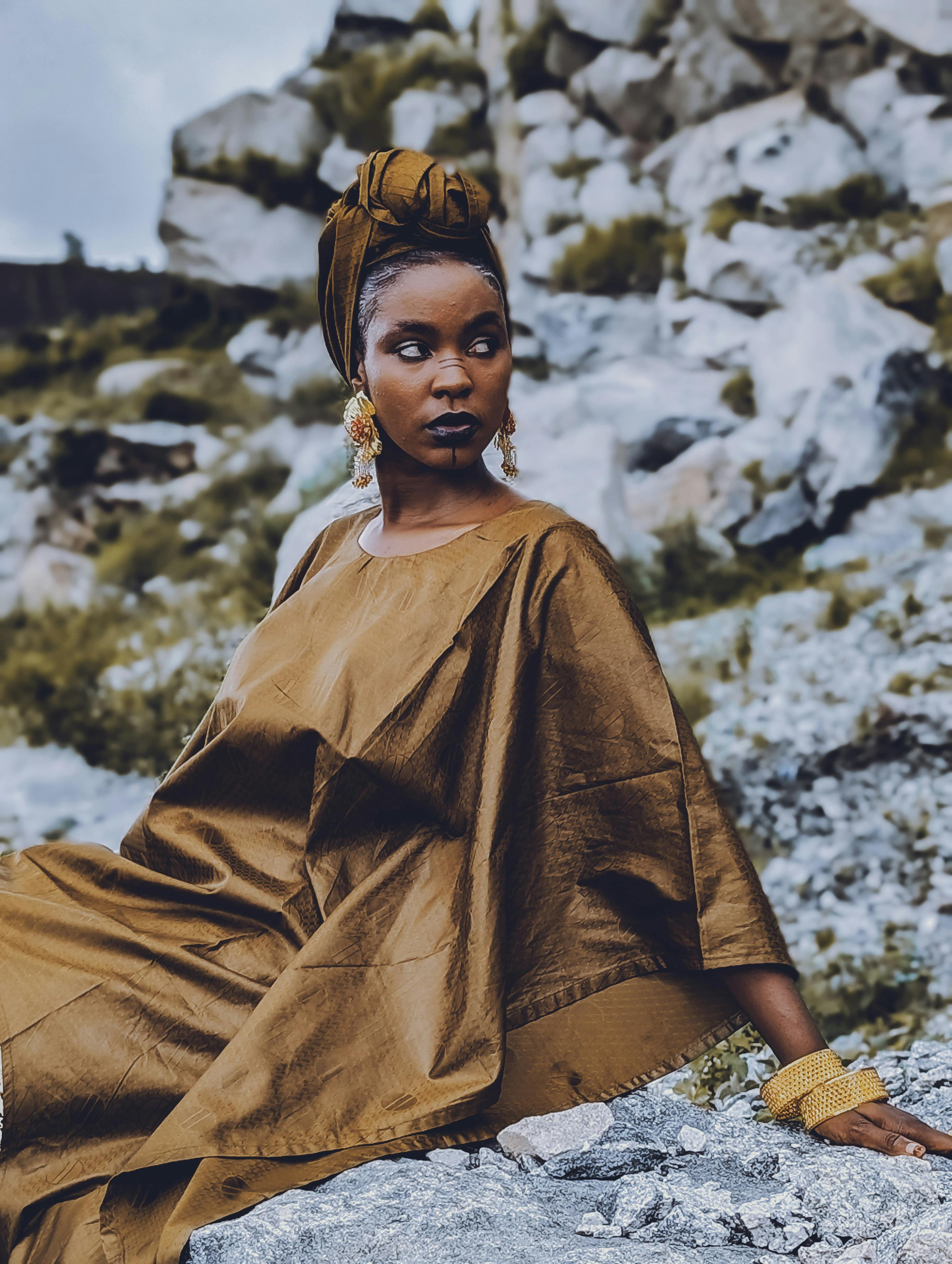 在阿布贾一处户外岩石背景前，一位身穿尼日利亚传统服饰的妇女正坐着