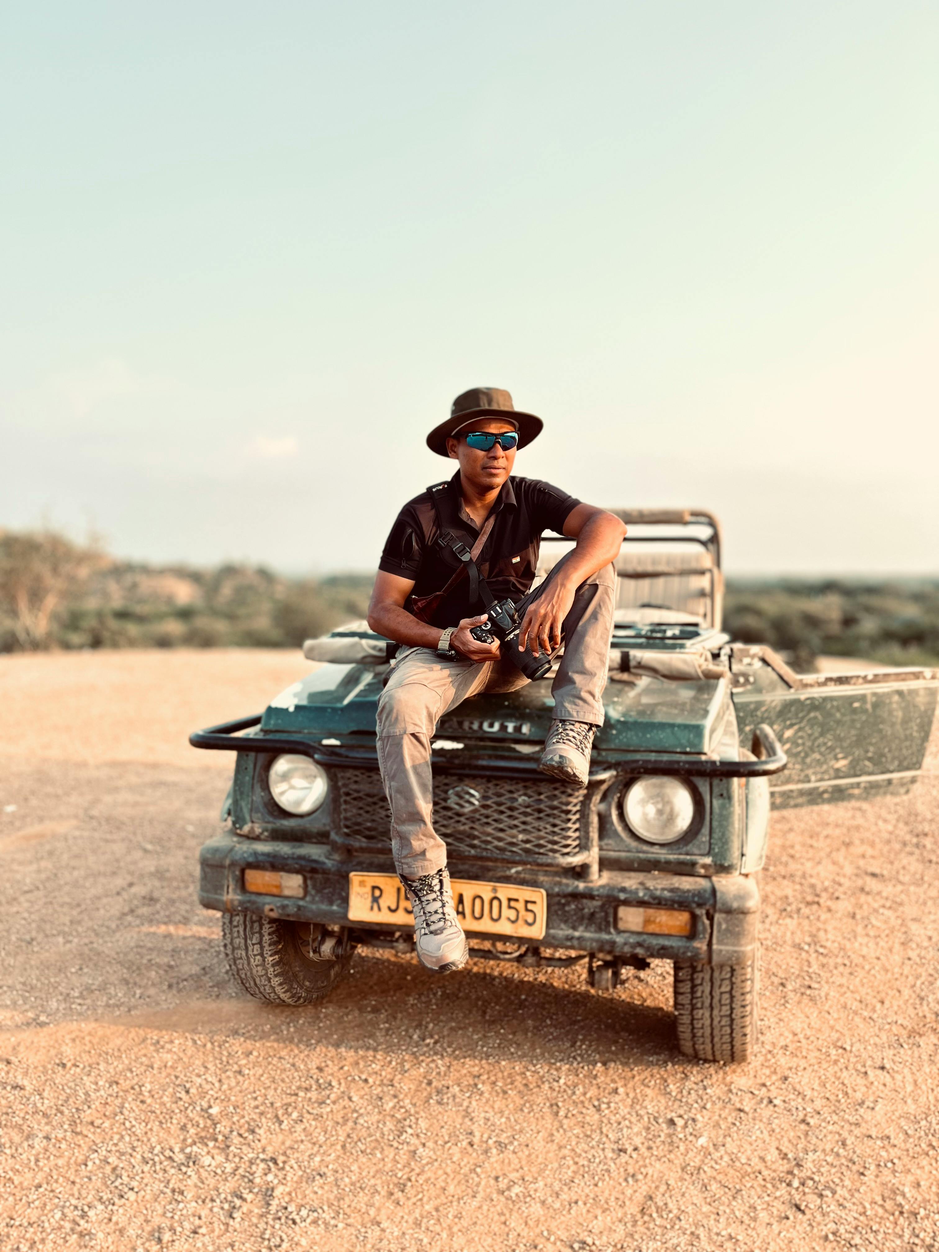 Man in hat and sunglasses sitting on jeep in arid terrain, ready for adventure.