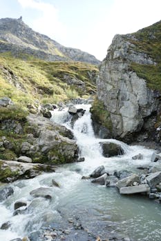 A tranquil mountain waterfall flows through a rocky landscape under a bright sky.