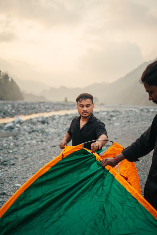 Free Two young men assembling a green and orange tent by a mountain river at sunset. Stock Photo