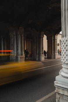 A yellow taxi blurs by as it passes under ornate stone arches in an urban setting, adding dynamic motion.