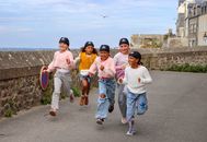 Group of children running happily wearing pirate hats on a seaside pathway.