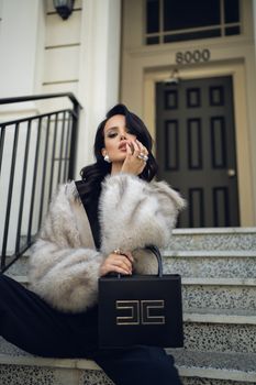 Fashionable woman in fur coat poses with luxury handbag on city steps.