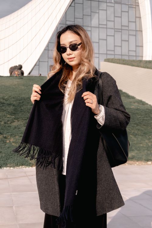 Free Stylish woman wearing sunglasses and coat with scarf standing outdoors by modern architectural building. Stock Photo