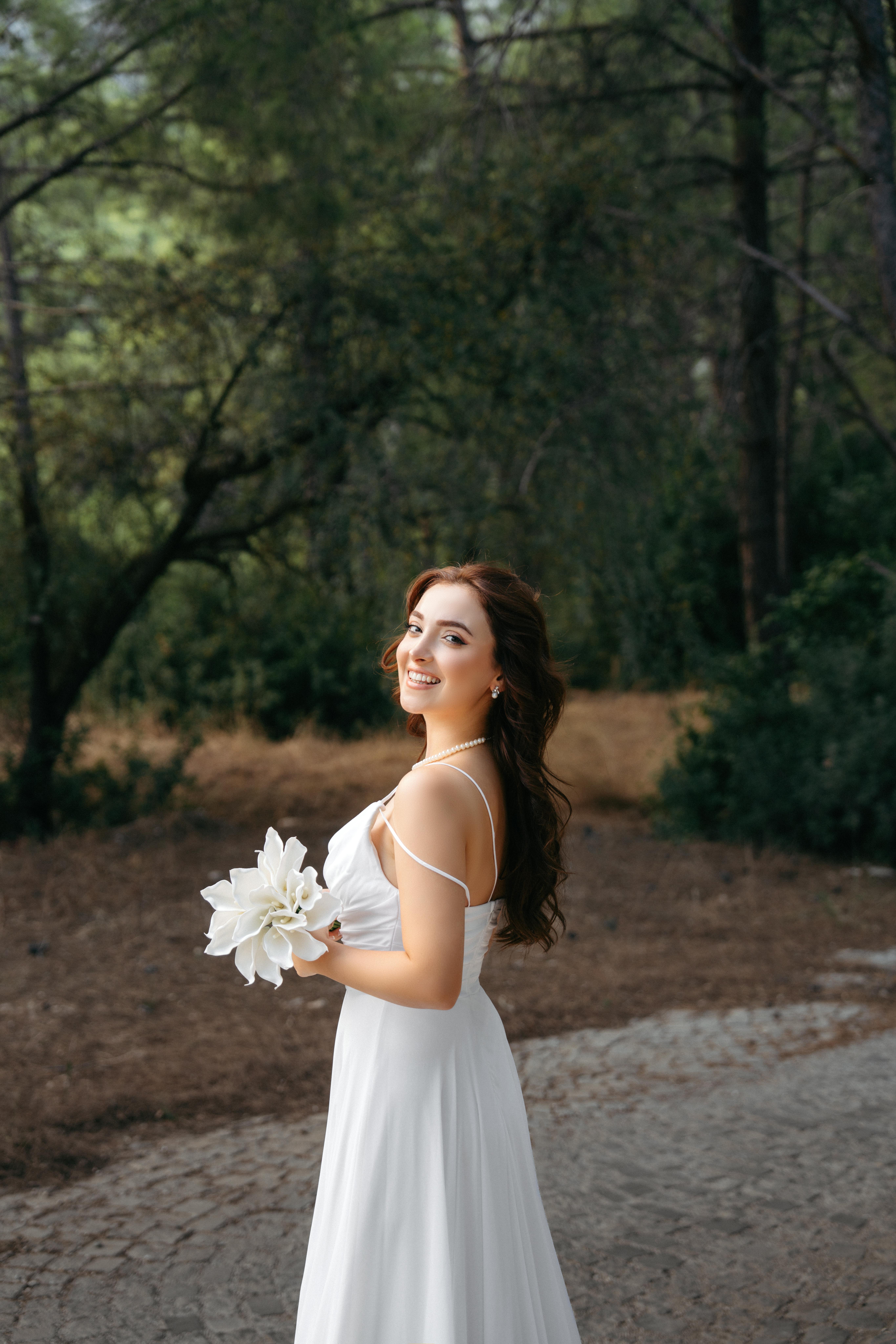 Elegant bride in white dress holding flowers in a serene Antalya forest setting.