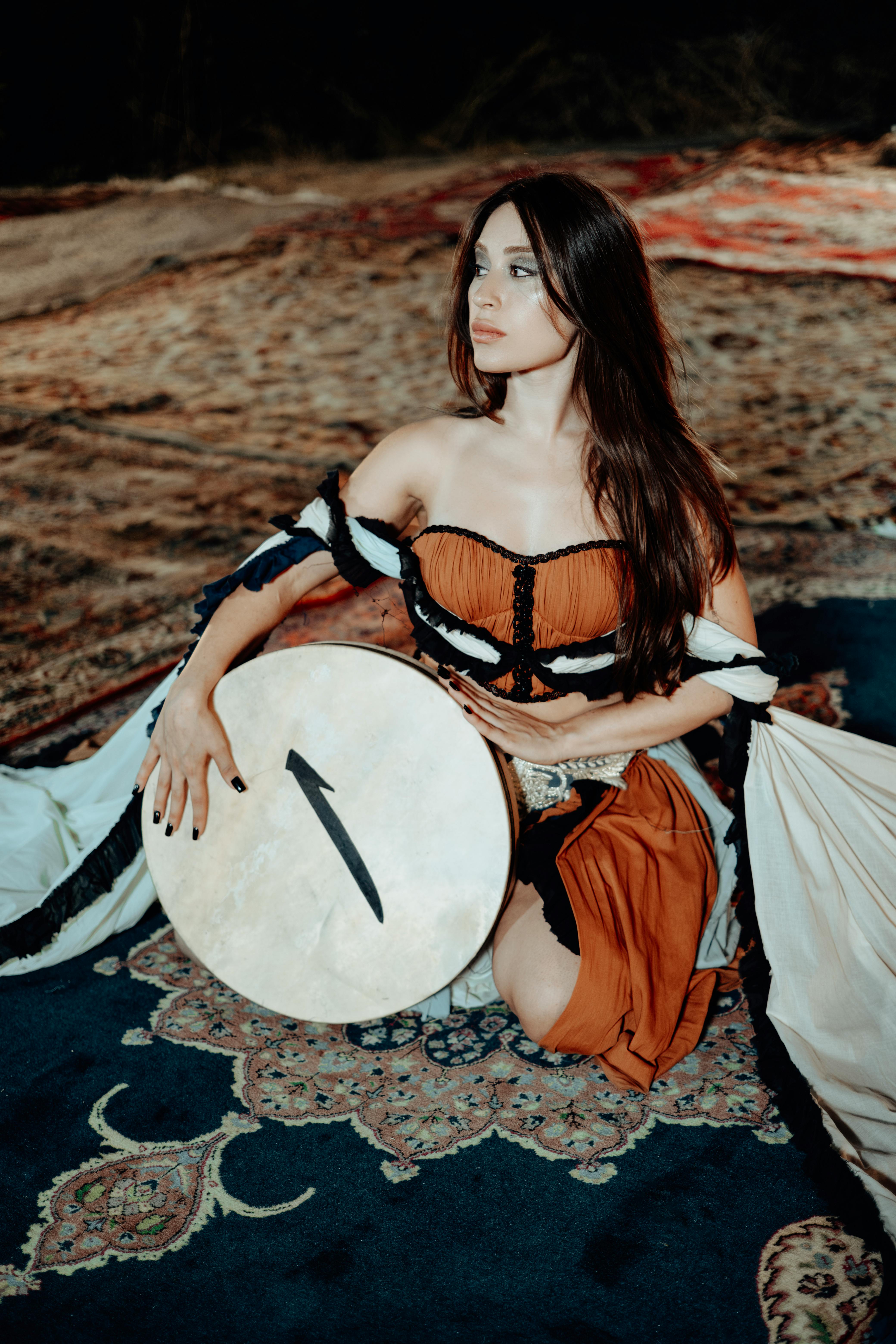 Woman in traditional attire holding a drum on patterned rugs in ambient setting.
