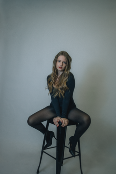A stylish woman poses confidently in a black outfit, seated on a stool in a studio setting.