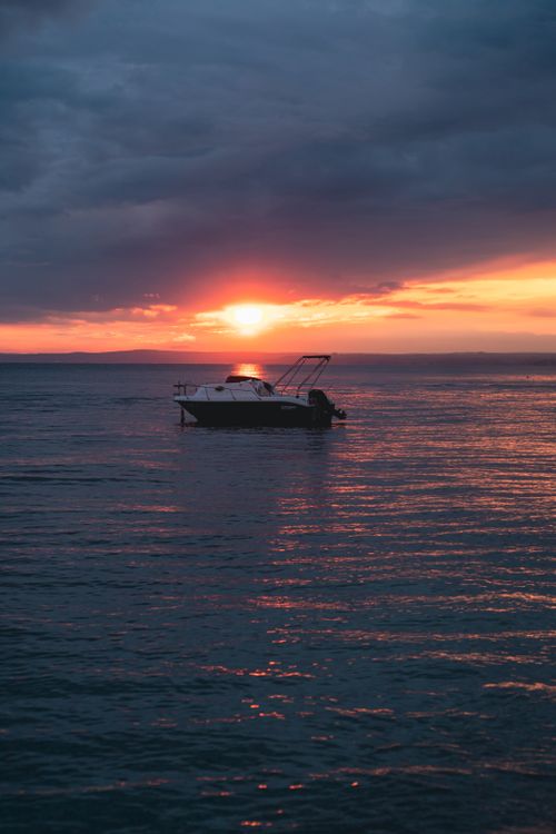 Free Serene sunset view with a solitary boat on calm waters under dramatic clouds. Perfect for relaxation themes. Stock Photo