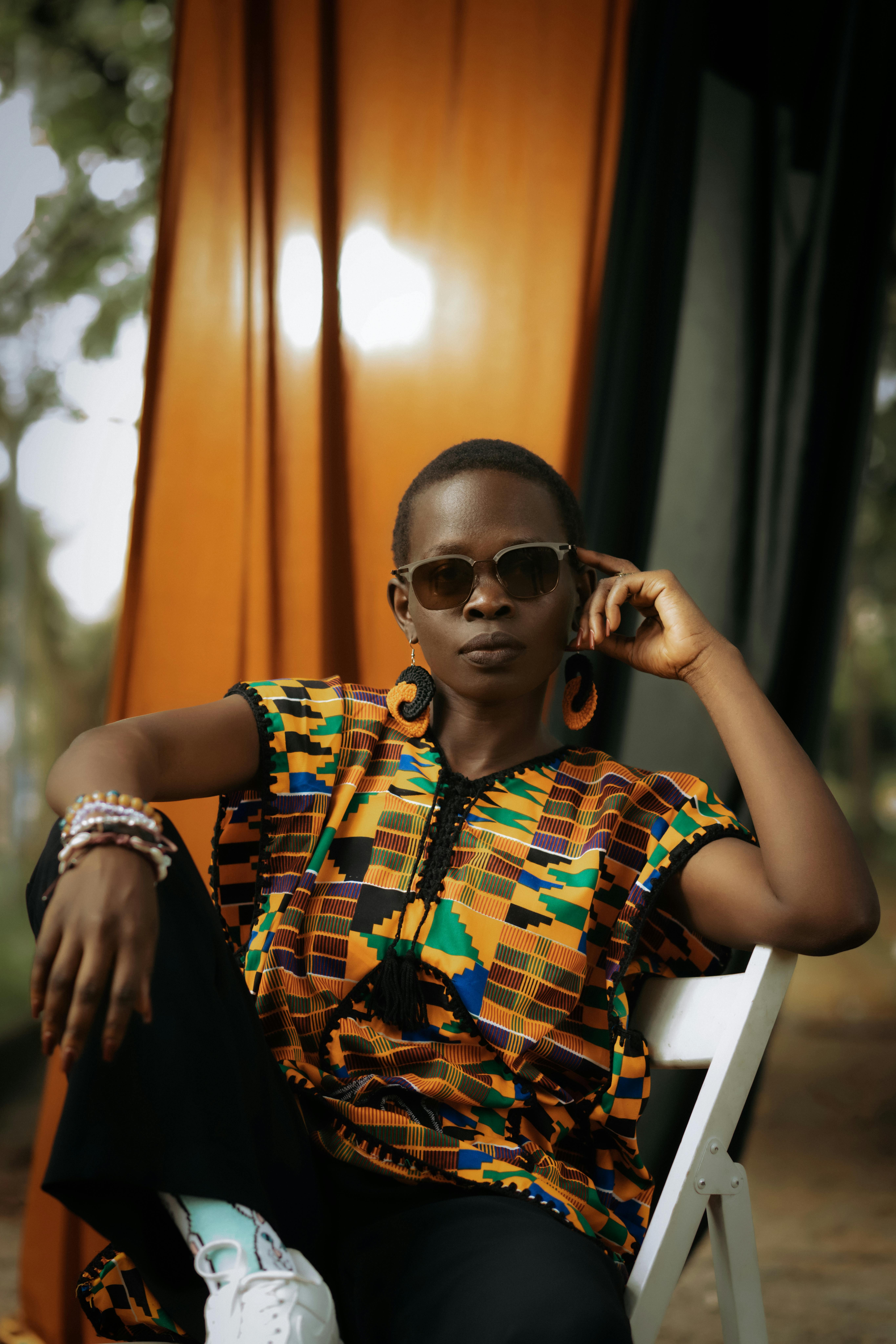 Stylish African woman wearing sunglasses and colorful Kente shirt, seated outdoors.