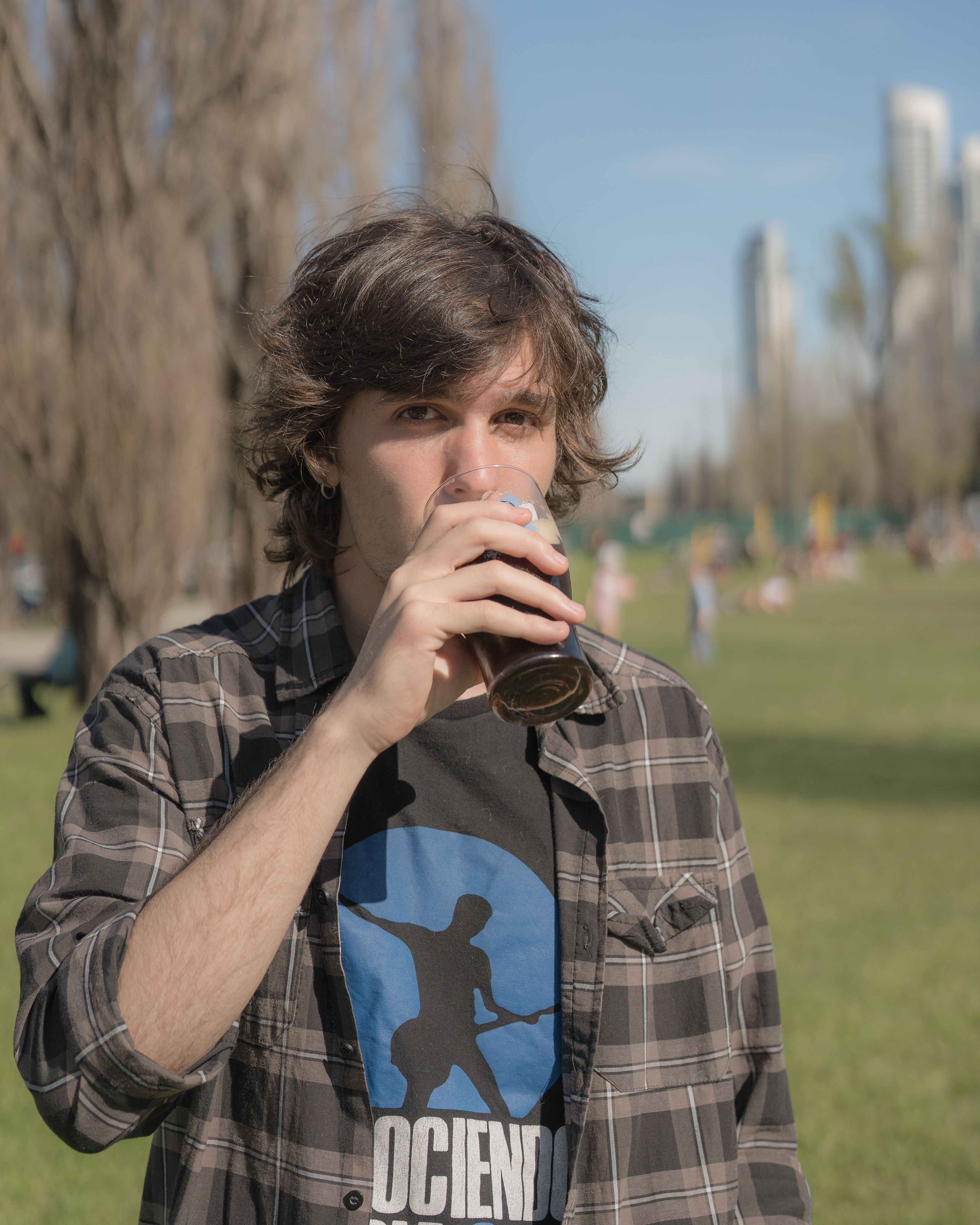 A young man drinks in a Buenos Aires park on a clear day, with skyscrapers in the background.