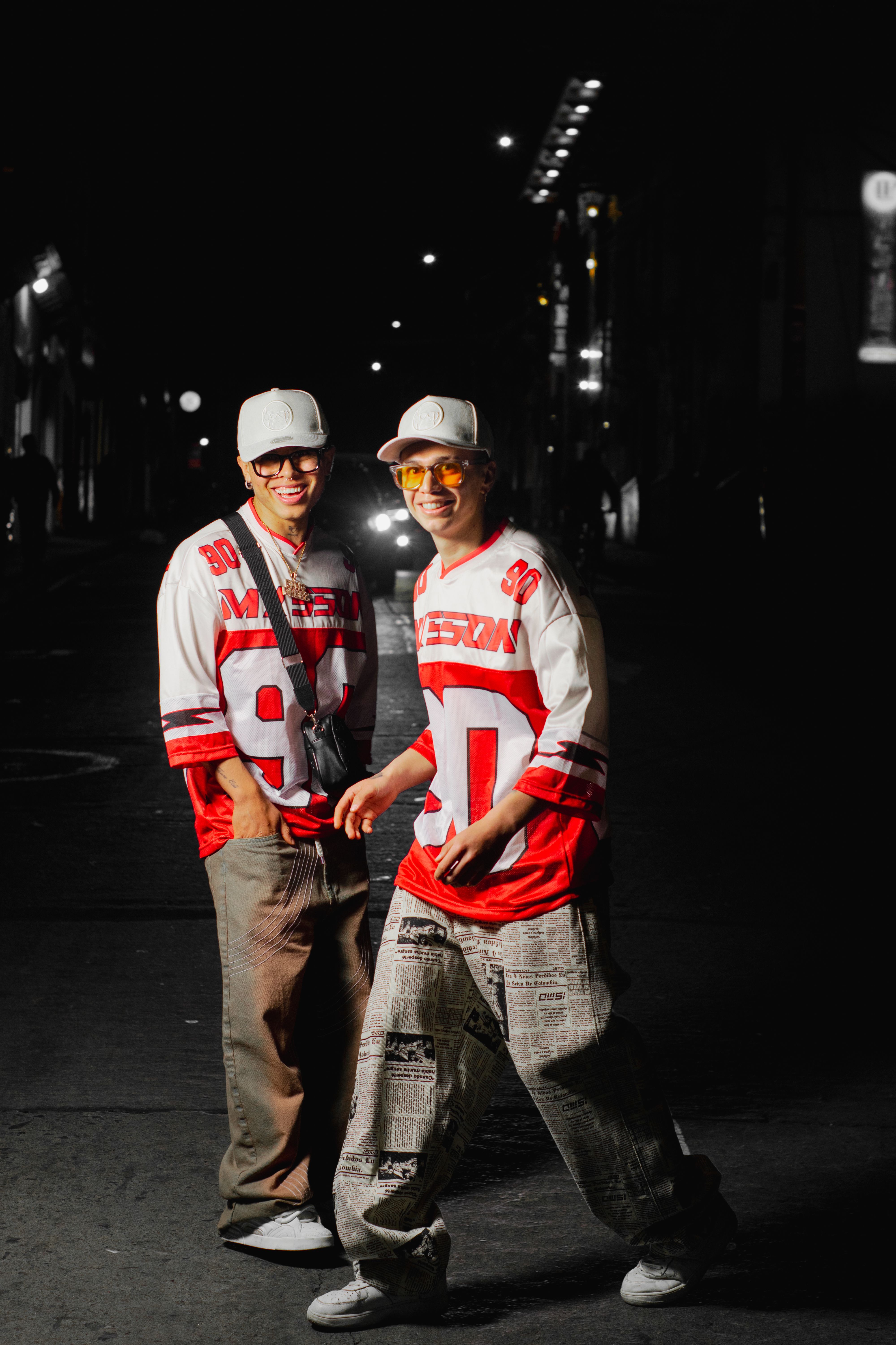 Two young men pose in matching sporty outfits on a dimly lit street in Colombia, showcasing urban fashion.