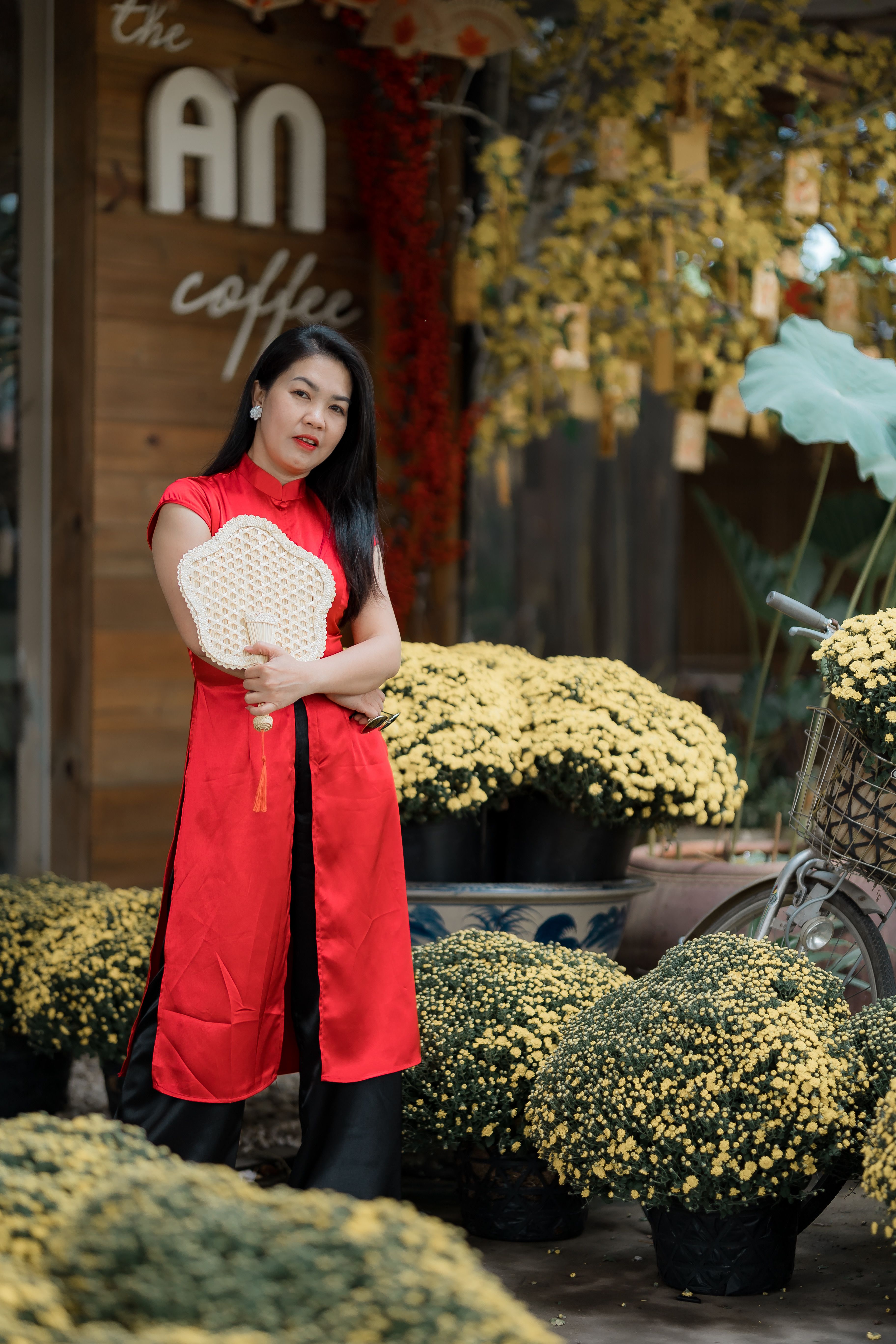 Stylish woman in traditional red dress with fan poses amid vibrant yellow flowers outside cafe.