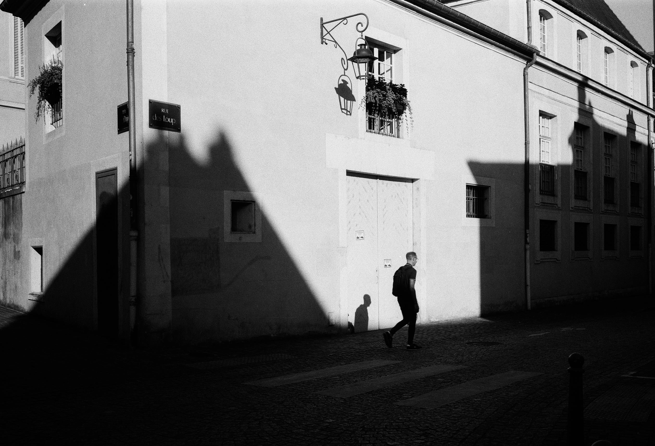 Silhouetted man walks past historic building in Nancy, France, casting long shadows.