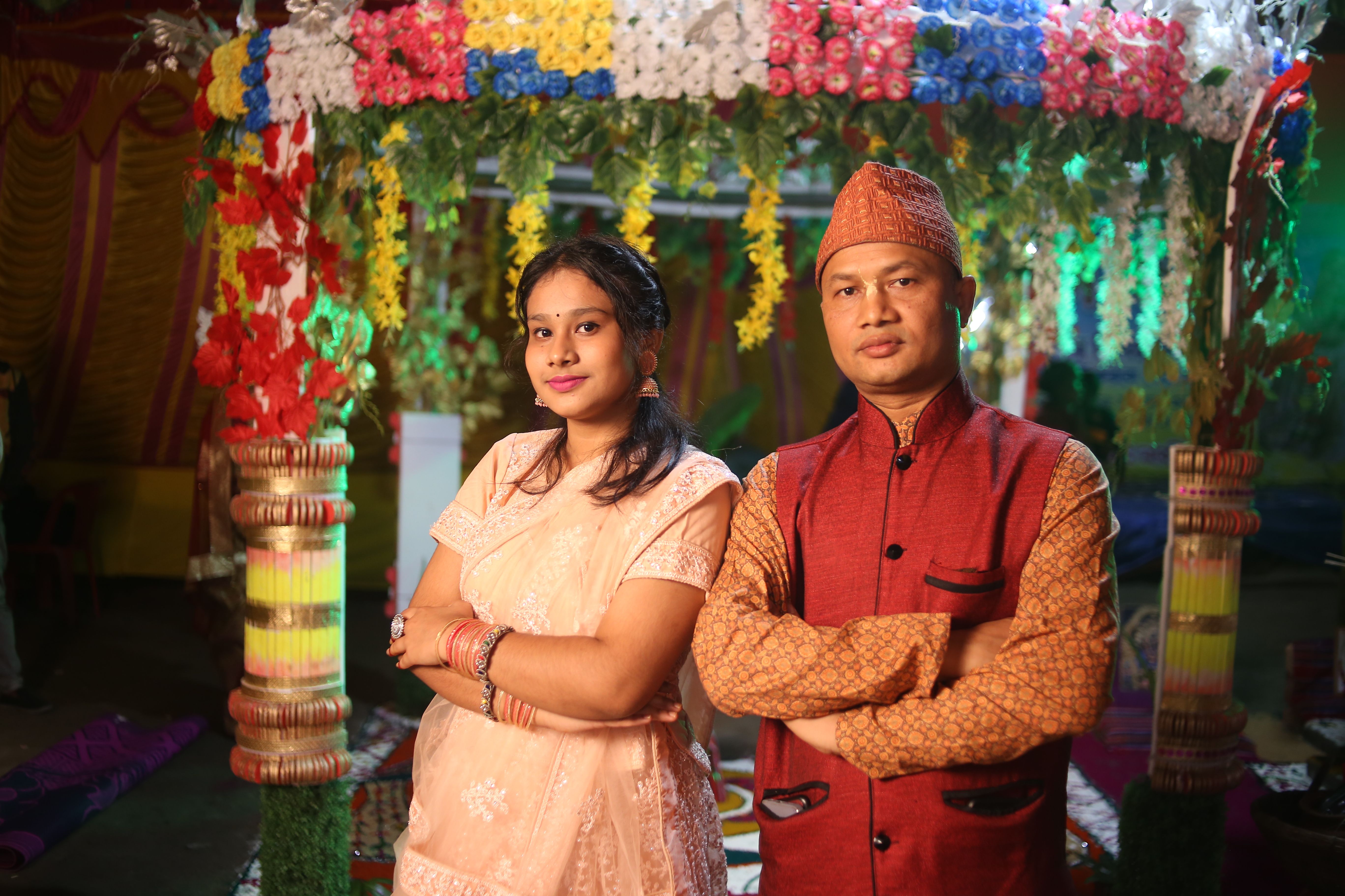 Couple in traditional attire standing indoors under colorful floral decorations during a festive event.