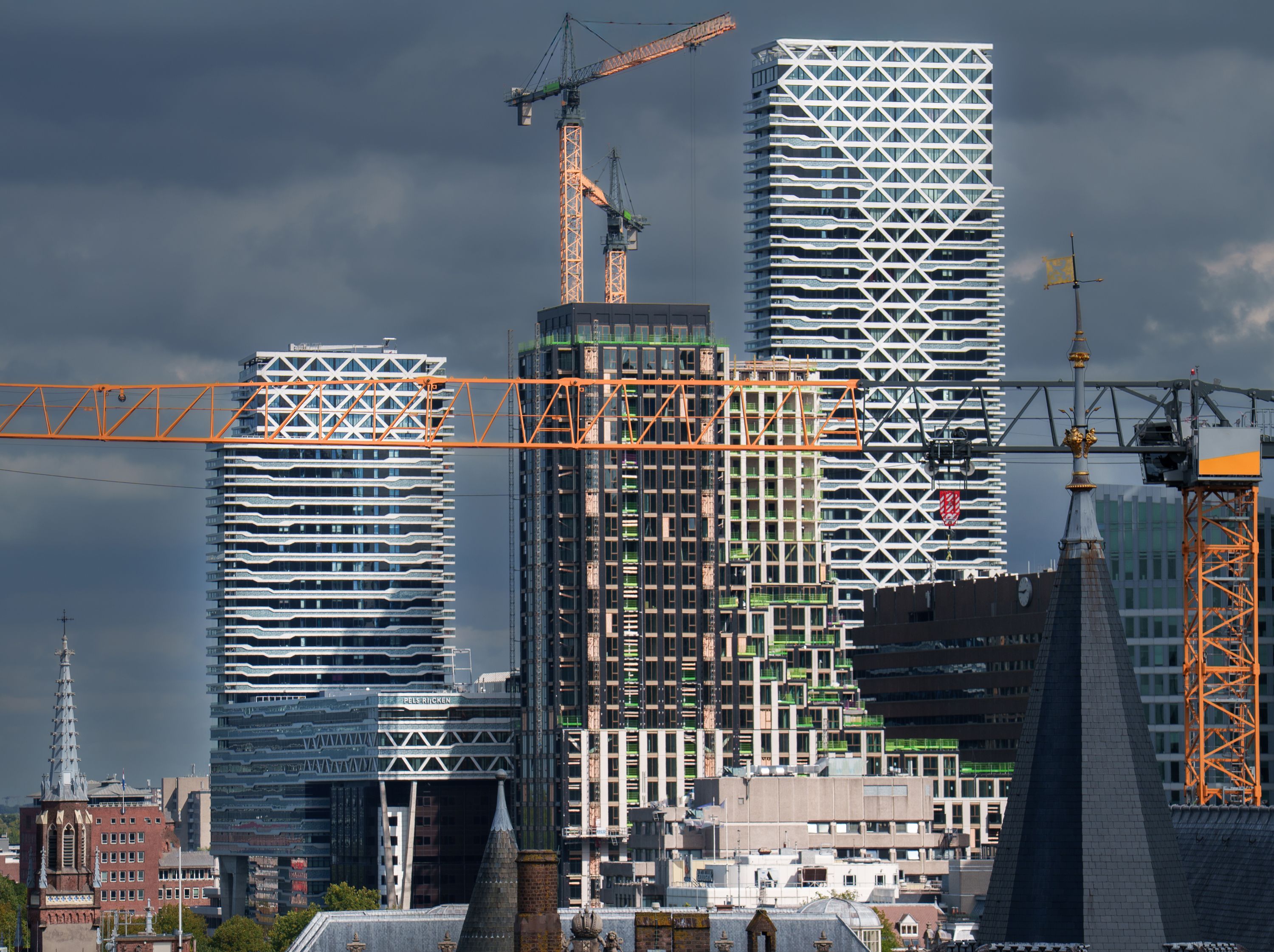 Construction cranes rise among modern high-rise buildings beneath dark clouds