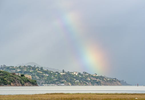 Vibrant rainbow arcs over a scenic coastal hillside town, reflecting a serene and colorful landscape.