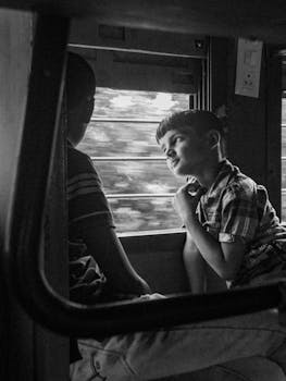 Black and white image capturing two children sitting by a train window, with one gazing thoughtfully outside.