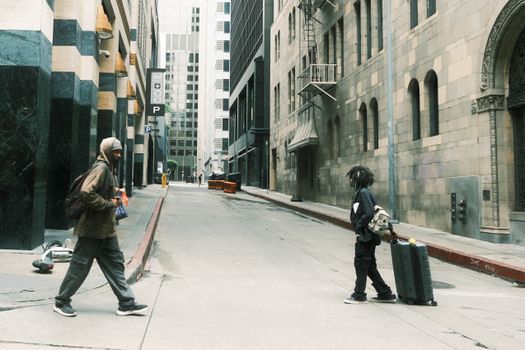 Two pedestrians crossing a street lined with tall buildings in downtown Los Angeles.