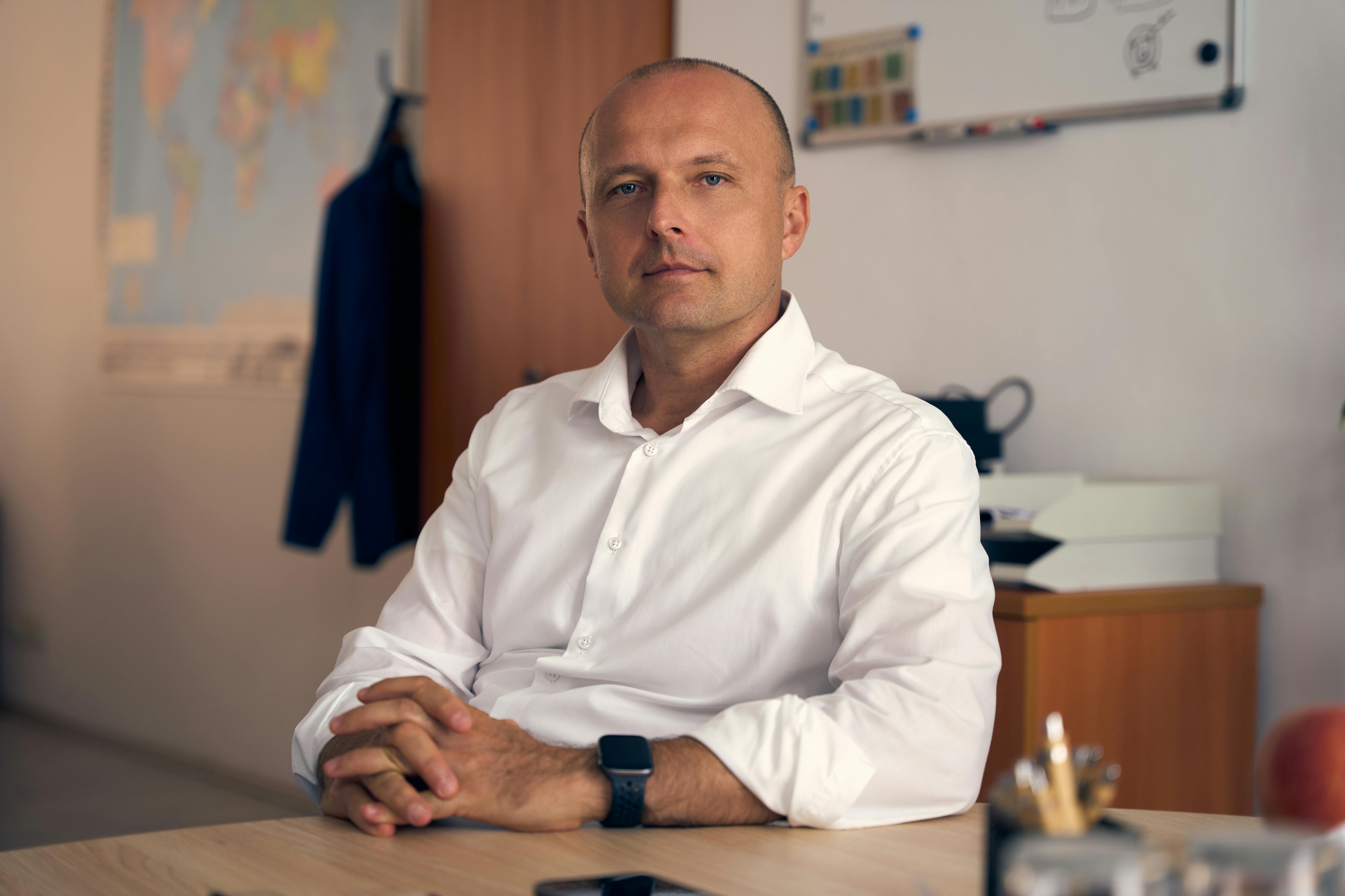 A confident adult man in a white shirt sitting in a well-lit office with a thoughtful expression.