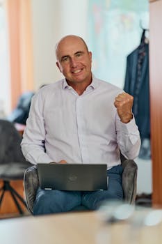 Smiling businessman in an office holding a laptop, expressing confidence and success.