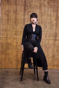 Tattooed woman in goth attire sits confidently on a stool, exuding style.