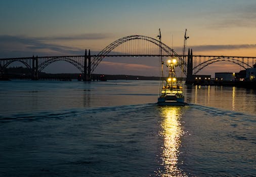 A fishing boat sails under an iconic bridge at sunrise, reflections on water.