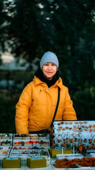 Smiling woman in yellow jacket selling handmade jewelry at an outdoor market stall.
