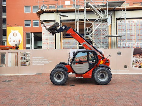 A red telehandler parked at a construction site with scaffolding and building materials in the background.