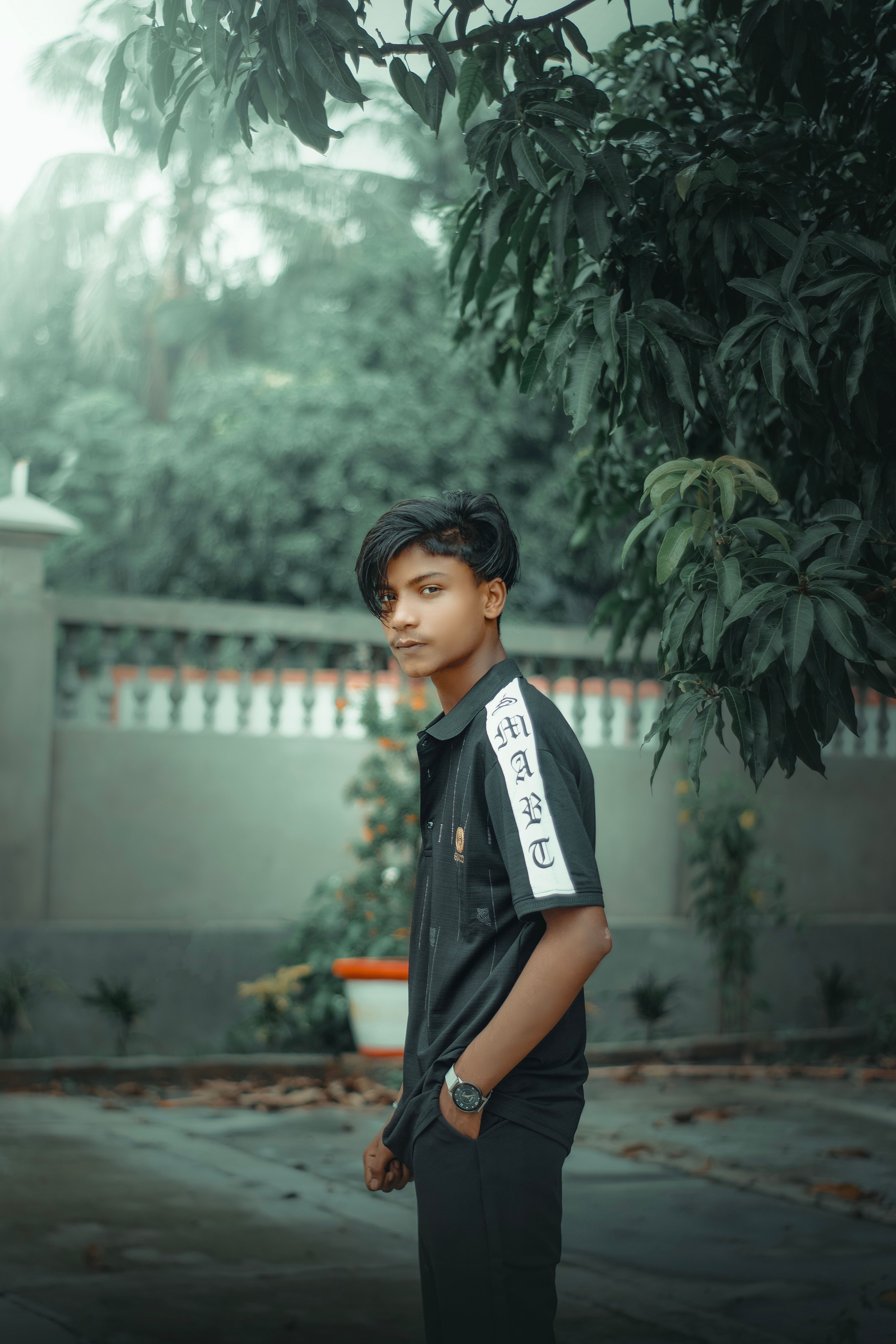 Portrait of a young man posing outdoors under a tree in a village setting.