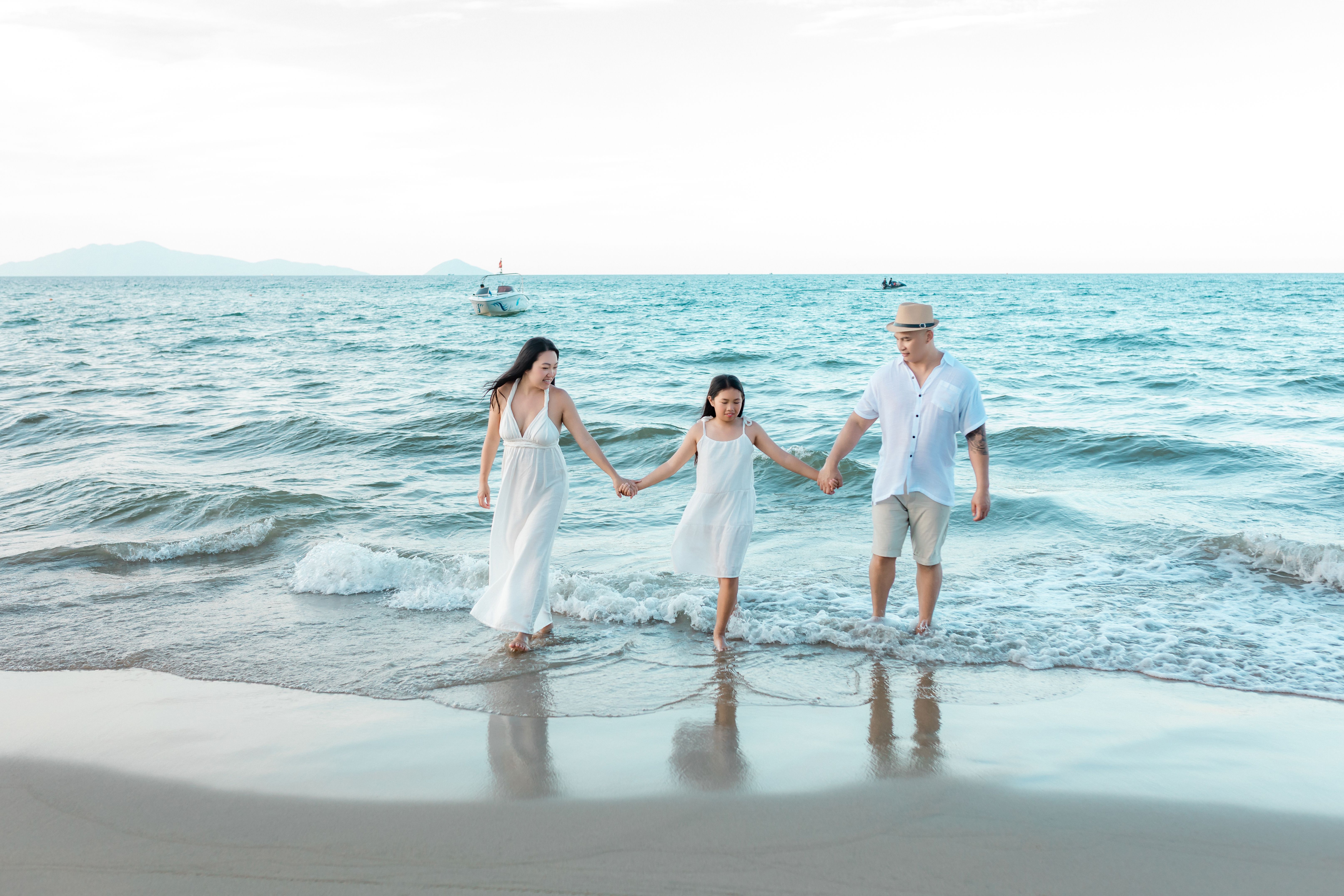 A family walking hand-in-hand in the surf at the beach, wearing light-colored summer clothing.