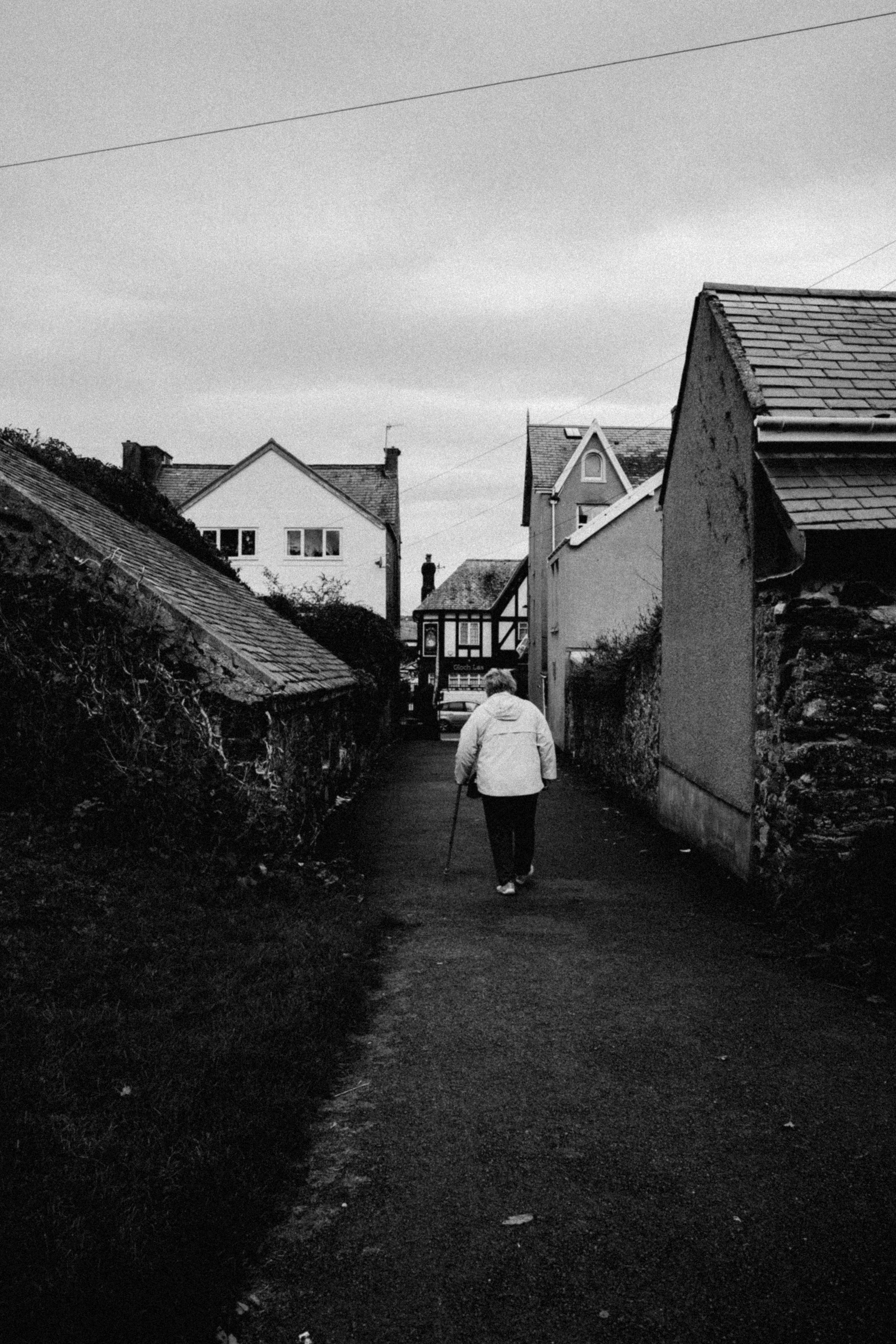 A senior woman walks down a narrow street in Conwy, Wales, embodying a serene moment in black and white.