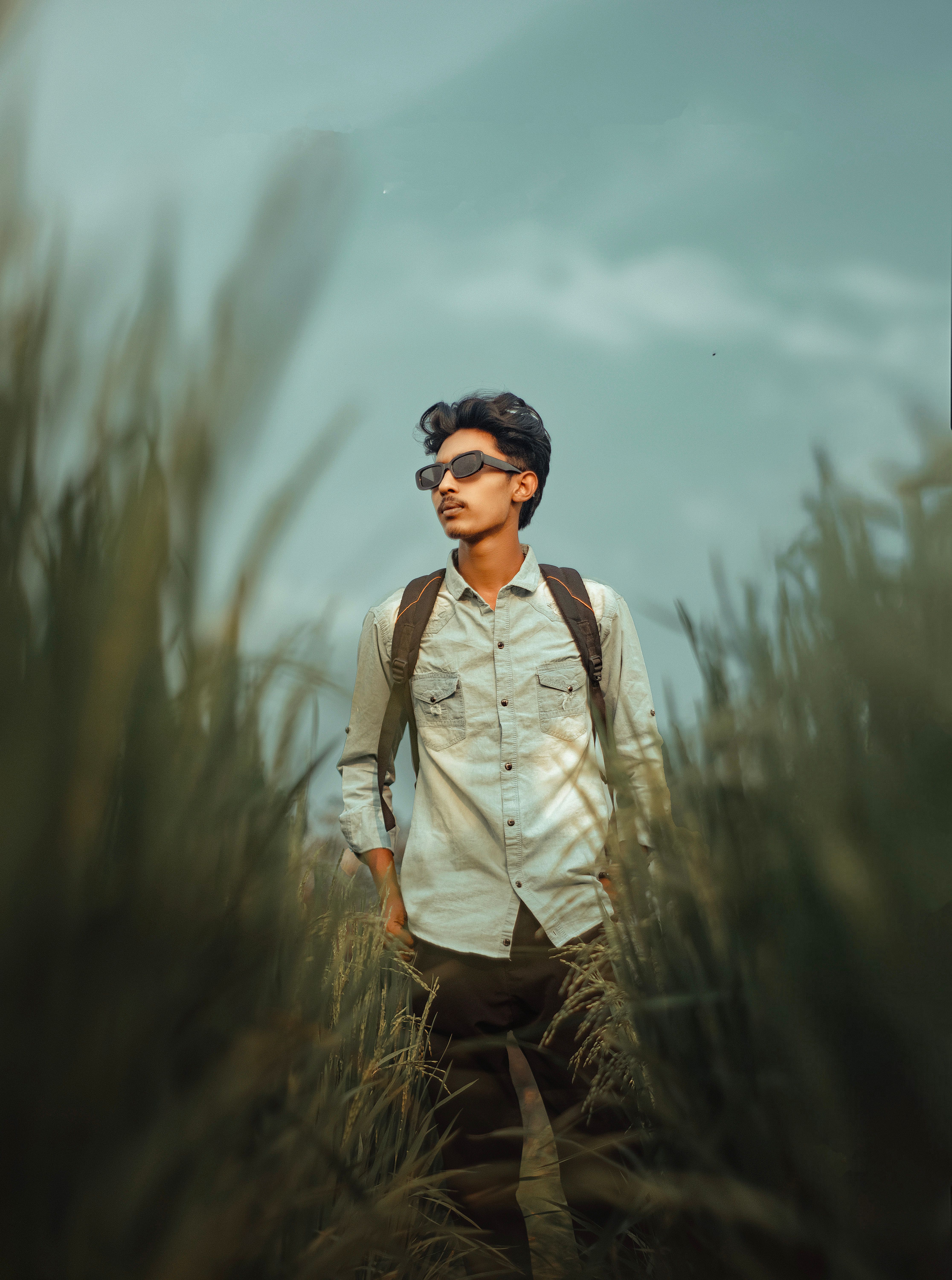 Stylish young man with backpack in a field wearing sunglasses, exuding adventure.